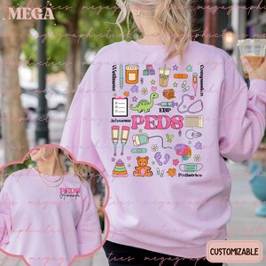 K&ouml;nnte beinhalten: Rosafarbener Pullover mit einem bunten Grafikdesign aus medizinischen Symbolen und dem Wort "PEDS" in der Mitte. Die Grafik enth&auml;lt ein Stethoskop, ein Thermometer, eine Spritze, einen Verband, einen Teddyb&auml;r, einen Dinosaurier und andere medizinische Symbole. Der Text "Advocate", "EBP", "Wellness", "Compassion" und "Pediatrics" ist ebenfalls in der Grafik enthalten.