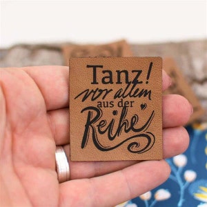 May include: A small, square, brown leather tag with the words "Tanz! vor allem aus der Reihe" in black script. The tag is held in a person's hand, with a silver ring visible. The background is blurred.
