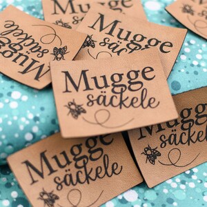Könnte beinhalten: Eine Ansammlung von hellbraunen Lederetiketten mit dem schwarzen Schriftzug "Mugge säckele". Jedes Etikett zeigt eine kleine Bienenillustration und eine geschwungene Linie. Die Etiketten liegen auf einem türkis-weiß gemusterten Stoff.