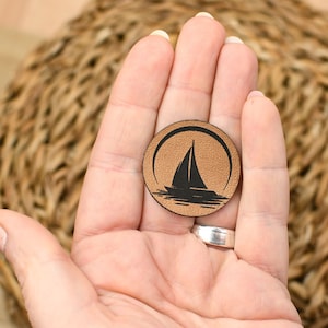 Könnte beinhalten: Ein braunes Leder-Patch mit einer schwarzen Silhouette eines Segelboots auf einer Wasserfläche. Das Patch ist rund und hat eine strukturierte Oberfläche.