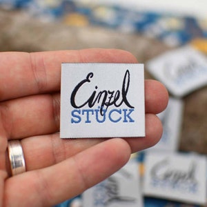 May include: A small, white fabric label with the word "Einzel" in black script above the word "STUCK" in blue block letters. The label is held in a hand, with other similar labels visible in the background.