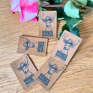 May include: A collection of rectangular, textured, tan leather tags with a deer illustration wearing a hat. The deer is in blue ink, and the text "WAIDHEIL MANNS" is printed below the deer. A pink rose and green leaves are in the background.