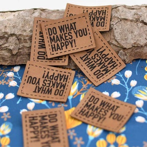 May include: A collection of brown leather patches with the text "Do what makes you happy!" printed in black. The patches have a stitched border and are scattered on a blue and white floral fabric.
