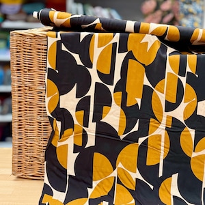 Könnte beinhalten: Stoffrolle mit einem auffälligen geometrischen Muster in Schwarz, Senfgelb und Creme. Der Stoff ist über einem Weidenkorb drapiert und zeigt das sich wiederholende abstrakte Design. Geeignet für verschiedene Nähprojekte.
