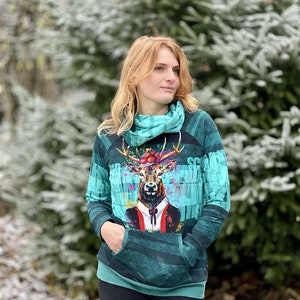 Könnte beinhalten: Ein teal- und grünfarbener Kapuzenpullover mit einem Hirsch-Grafik, der einen Anzug und eine Krawatte trägt. Der Pullover hat einen Rollkragen und eine Kängurutasche. Der Hintergrund ist eine verschneite Szene im Freien.