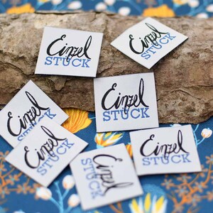May include: A collection of small, square white fabric labels with the German words "Einzel Stuck" printed in black script and blue block letters. The labels are scattered on a textured wooden surface.