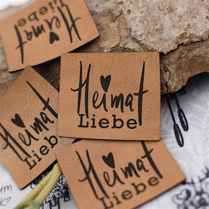 Könnte beinhalten: Mehrere quadratische, hellbraune Lederetiketten mit dem schwarzen Schriftzug "Heimat Liebe". Ein kleines schwarzes Herz befindet sich über dem Wort "Liebe". Die Etiketten liegen auf einer strukturierten Oberfläche.