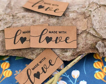 May include: Tan rectangular labels with the words "MADE WITH love" in black script. A black heart replaces the "o" in love. The labels are arranged on a textured, light brown surface and a blue floral fabric.