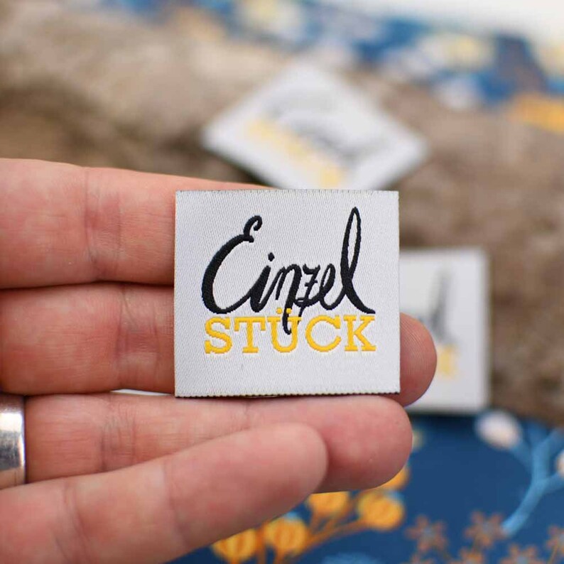 May include: White woven fabric label with black cursive text "Einzel" and yellow text "STUCK".