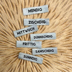 Peut inclure: Sept petites étiquettes rectangulaires grises avec du texte noir indiquant les jours de la semaine en allemand : MENDIG, ZISCHDIG, MITTWOCH, DUNNSCHDIG, FRITTIG, SAMSCHDIG et SUNNDIG. Les étiquettes sont disposées sur une surface texturée brun clair.