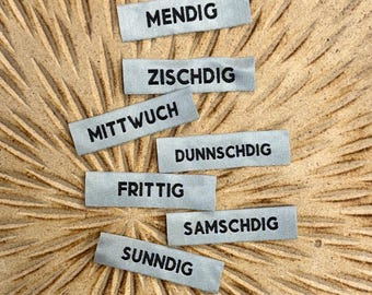May include: Seven small, rectangular grey fabric labels with black text spelling out the days of the week in German: MENDIG, ZISCHDIG, MITTWOCH, DUNNSCHDIG, FRITTIG, SAMSCHDIG, and SUNNDIG. The labels are arranged on a textured, light brown surface.