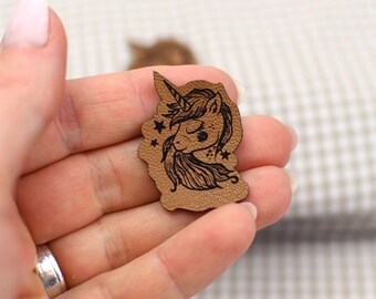 May include: A brown leather patch featuring a black illustration of a unicorn's head with a horn, flowing mane, and stars. The patch has a unique shape and is likely used for crafting or embellishment.