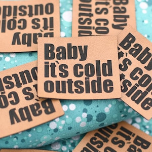 Könnte beinhalten: Nahaufnahme von mehreren hellbraunen Lederetiketten mit dem schwarzen Aufdruck "Baby it's cold outside". Die Etiketten liegen auf einem türkisfarbenen Stoff mit weißen und silbernen Punkten.