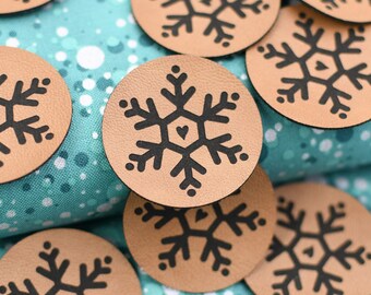 May include: A collection of round, tan leather patches with a black snowflake design. Each patch features a detailed snowflake with a heart in the center. The patches are scattered on a teal and white patterned fabric.