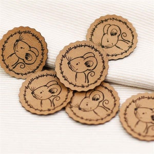 Könnte beinhalten: Sechs braune Leder-Patches mit einer genähten Kante und einer schwarzen Umrandung eines Mäusegesichts. Die Maus hat einen kleinen Schwanz und große Ohren.