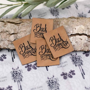 Könnte beinhalten: Vier quadratische, hellbraune Lederetiketten mit dem Schriftzug "Black Forest" in schwarzer Schrift und einer Berg- und Baumgrafik. Die Etiketten sind auf einem Holzstück angeordnet, mit einem gemusterten Stoff im Hintergrund.