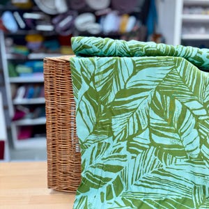 Könnte beinhalten: Ein geflochtener Weidenkorb enthält einen gefalteten Stoff mit einem tropischen Blättermuster. Der Stoff zeigt große grüne Blätter auf hellblauem Hintergrund. Der Korb ist aus braunem Weidengeflecht.