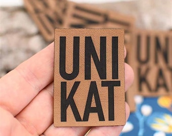May include: A rectangular, tan leather patch with the word "UNIKAT" printed in bold black letters. The patch is held in a hand, with other similar patches visible in the background. The leather has a textured appearance.