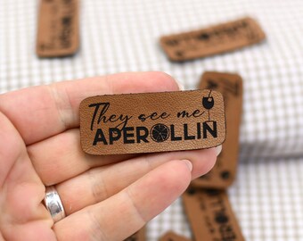 May include: Brown leather patch with the text "They see me Aperol-lin" in black lettering. A small orange slice and a glass of wine are also included in the design.