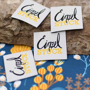May include: Four white fabric labels with black text that reads "Einzel Stuck" in a cursive font. The text is outlined in yellow.