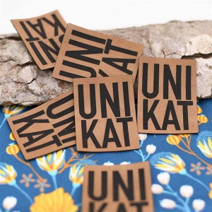 May include: Close-up of five brown leather patches with black text that reads "Unikat". The patches are arranged in a pile on a wooden surface.