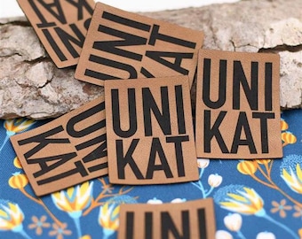 May include: Close-up of five brown leather patches with black text that reads "Unikat". The patches are arranged in a pile on a wooden surface.