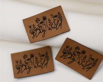 May include: Three rectangular brown leather tags with the words "stay wild" in a cursive font. Each tag features a black floral design above the text. The tags are arranged on a white fabric background.
