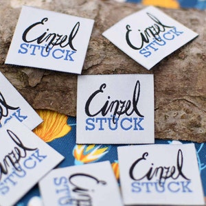 May include: Several white fabric labels with the word "Einzel" in a black script font above the word "STUCK" in blue block letters. The labels are square or diamond-shaped and are scattered on a wooden surface and a blue patterned fabric.