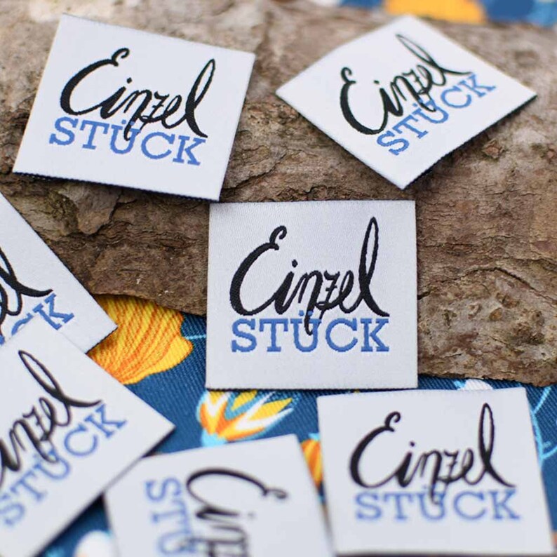 May include: Several white fabric labels with the word "Einzel" in a black script font above the word "STUCK" in blue block letters. The labels are square or diamond-shaped and are scattered on a wooden surface and a blue patterned fabric.