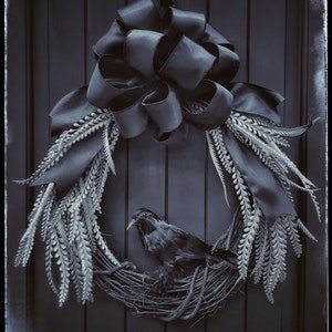 May include: A black crow sits in a wreath made of white twigs and a black satin bow. The wreath is hanging on a dark wooden door.