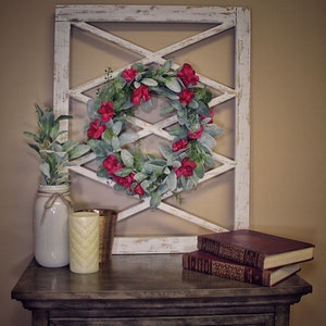 May include: A rustic, white-painted wooden wall decor with an X-pattern design, adorned with a wreath of red flowers and green leaves. A nightstand holds a mason jar with greenery, candles, and books, creating a cozy, farmhouse aesthetic.