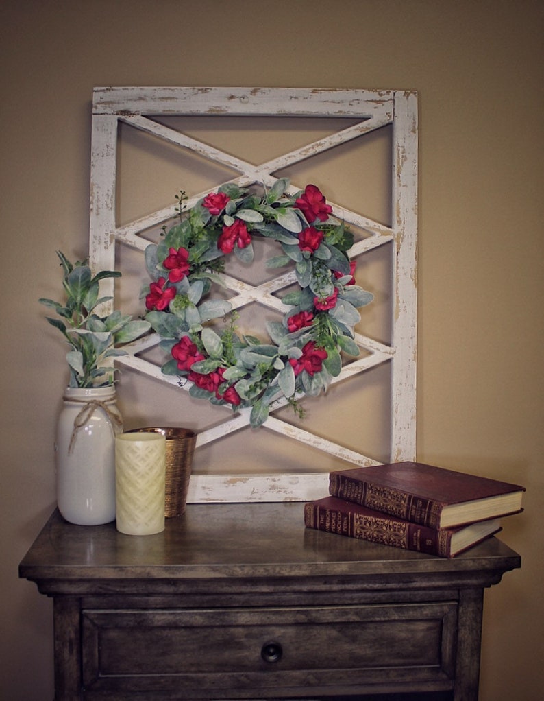 May include: A rustic, white-painted wooden wall decor with an X-pattern design, adorned with a wreath of red flowers and green leaves. A nightstand holds a mason jar with greenery, candles, and books, creating a cozy, farmhouse aesthetic.