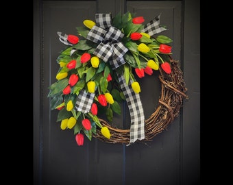 May include: A spring wreath featuring red and yellow tulips, green leaves, and a black and white checkered bow. The wreath is made of woven twigs and is designed for a front door.