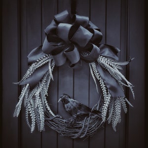 May include: A black crow sits in a wreath made of faux greenery and a black satin bow. The wreath is hanging on a dark wooden door.