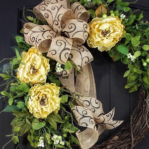 Pode incluir: Uma coroa de juta marrom com um grande la&ccedil;o e flores amarelas. O la&ccedil;o tem um padr&atilde;o de redemoinho marrom. A coroa &eacute; decorada com folhas verdes e bagas brancas.