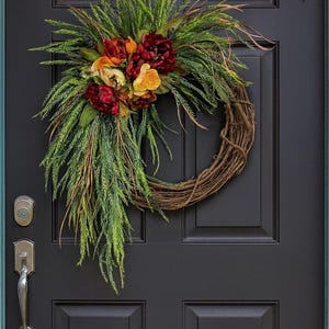 Puede incluir: Una corona decorativa con una base de vid natural, adornada con vibrantes flores rojas, amarillas y naranjas, y un exuberante follaje verde. La corona se exhibe en una puerta de entrada negra.