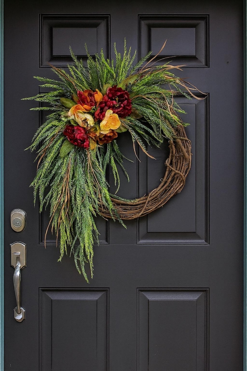 Puede incluir: Una corona decorativa con una base de vid natural, adornada con vibrantes flores rojas, amarillas y naranjas, y un exuberante follaje verde. La corona se exhibe en una puerta de entrada negra.