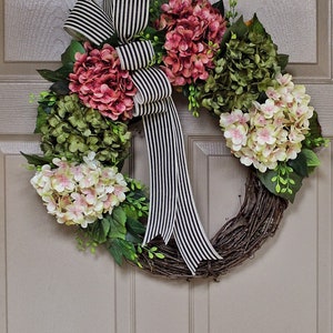 Pu&ograve; includere: Una ghirlanda di vite con fiori di ortensia rosa, bianchi e verdi. Un nastro a righe bianche e nere &egrave; legato in un fiocco al centro della ghirlanda.
