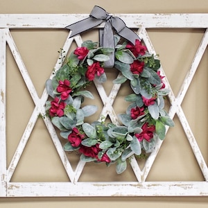 May include: A white, distressed wooden wall decor piece with a diamond-shaped lattice design. A wreath of red flowers and green foliage is centered within the diamond. A black and white striped bow is at the top.