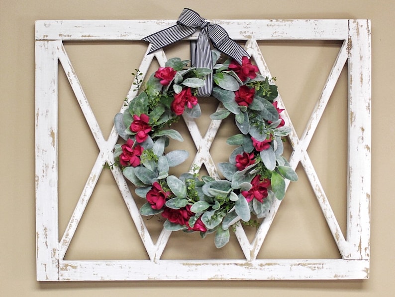 May include: A white, distressed wooden wall decor piece with a diamond-shaped lattice design. A wreath of red flowers and green foliage is centered within the diamond. A black and white striped bow is at the top.