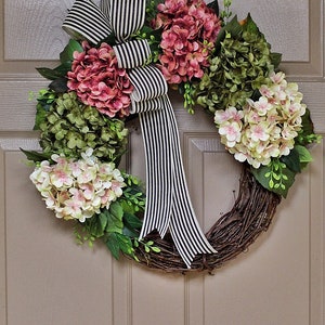 Pu&ograve; includere: Una ghirlanda di vite con fiori di ortensia rosa, bianchi e verdi. Un nastro a righe bianche e nere &egrave; legato in un fiocco sulla ghirlanda.