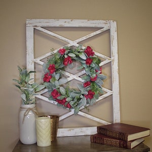 May include: A decorative arrangement featuring a white distressed window frame with a red flower wreath. A white jar with greenery, a gold candle holder, a cream candle, and two books are on a wooden table.