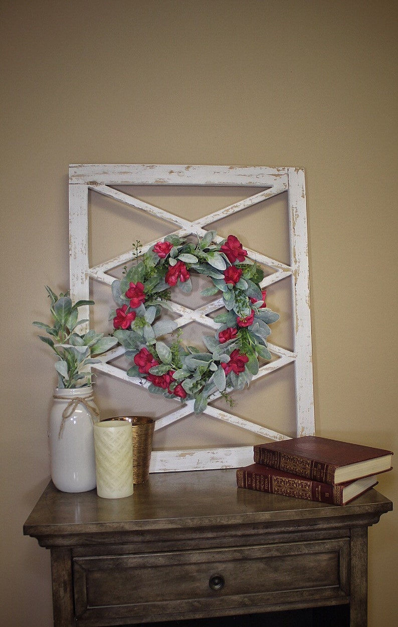 May include: A decorative arrangement featuring a white distressed window frame with a red flower wreath. A white jar with greenery, a gold candle holder, a cream candle, and two books are on a wooden table.