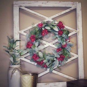 May include: A white distressed wooden wall decor with an X-shaped design, adorned with a floral wreath of red flowers and green leaves. A white mason jar with greenery, a candle, and books complete the display.