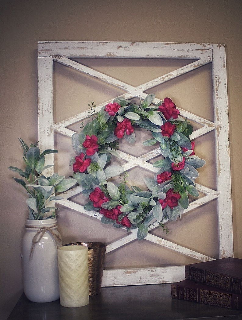 May include: A white distressed wooden wall decor with an X-shaped design, adorned with a floral wreath of red flowers and green leaves. A white mason jar with greenery, a candle, and books complete the display.