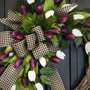 May include: A grapevine wreath with a black and white checkered ribbon bow and a cascade of white and burgundy tulips.