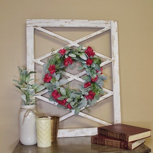 May include: A decorative farmhouse-style display featuring a white distressed wooden lattice, a floral wreath with red flowers, and a mason jar with greenery. Additional decor includes candles and books on a wooden table.