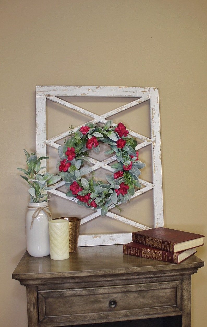 May include: A decorative farmhouse-style display featuring a white distressed wooden lattice, a floral wreath with red flowers, and a mason jar with greenery. Additional decor includes candles and books on a wooden table.