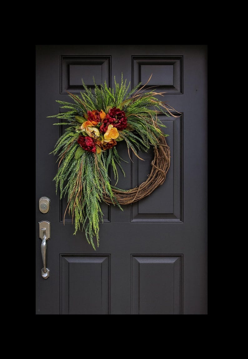 Puede incluir: Una corona decorativa hecha de ramitas trenzadas, adornada con follaje verde y flores artificiales coloridas en tonos rojos, naranjas y amarillos. La corona cuelga de una puerta negra con manija y cerradura plateadas.