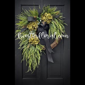 May include: A green and black wreath with a black ribbon bow. The wreath is made of greenery and has a cluster of green hydrangeas. The wreath is hanging on a black door. The text "fivestarfloral.com" is visible in the image.